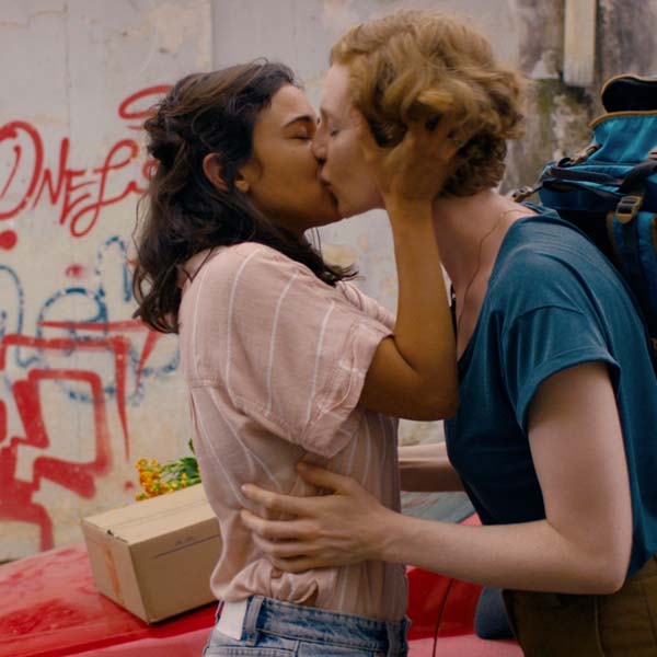 Two women kissing in a street scene next to a car.