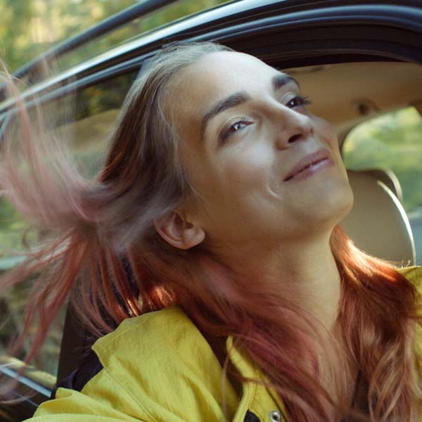 A trans girl hanging her head outside of a car window smiling as the breeze blows through her hair.