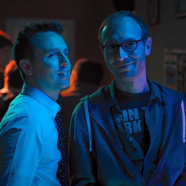 Two men stand in a bar, washed over with a blue light