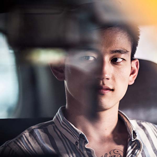 An asian man wearing a striped shirt sits in a car waiting looking into the distance