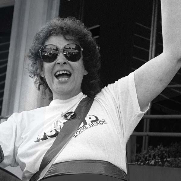 A trans woman with short curly hair, wearing sunglasses, stands with her fist raised in solidarity