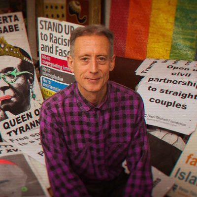 Portrait or Peter Tatchall surrounded by protest signage and a pride flag.