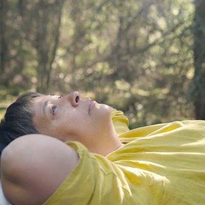 A woman in a yellow top laying on the ground—surrounded by trees—looking at up at the sky.