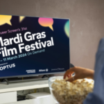 A TV screen that has the MGFF24 logo and speech bubble design on it. Someone is sitting in front of the tv with popcorn.
