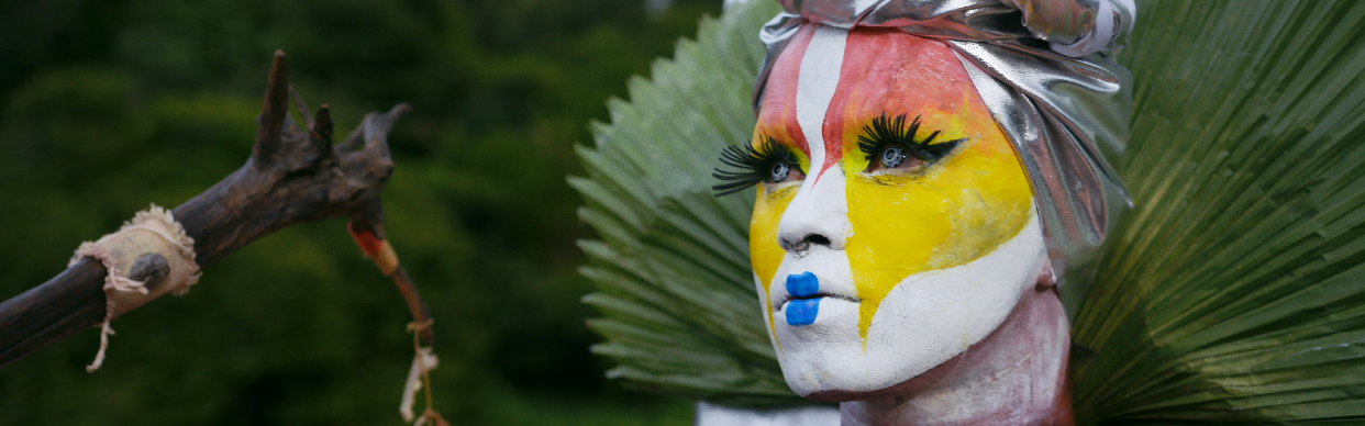 Uýra dressed for their performance. Their face painted in white, yellow and red with blue lips.