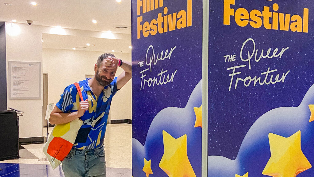 A person wearing a blue button up leans against a purple sign that says "Film Festival Queer Frontier". They have short brown hair with a beard, and they are smiling.