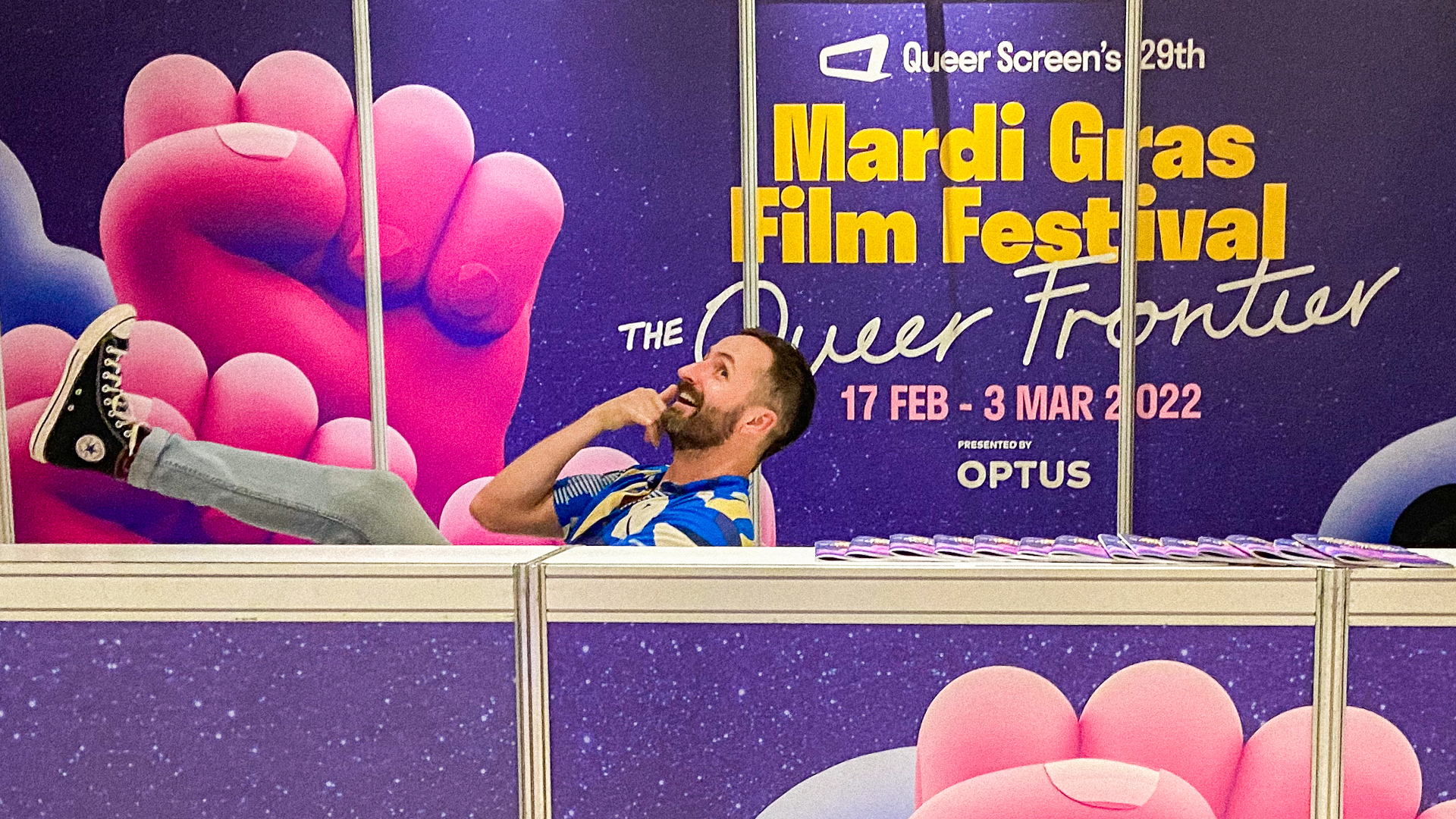 A person wearing a blue button up is sitting on a chair, resting their legs up on the desk, talking on the phone while smiling. The desk and backdrop are purple, and the backdrop says "Mardi Gras Film Festival Queer Frontier".