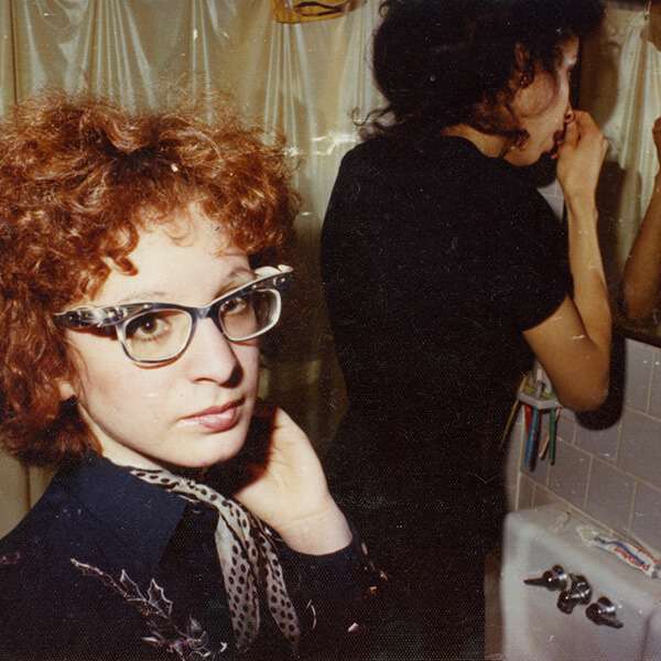 Two women standing in bathroom, putting on make up and fixing their hair. One of the women is wearing glasses and staring into the camera.