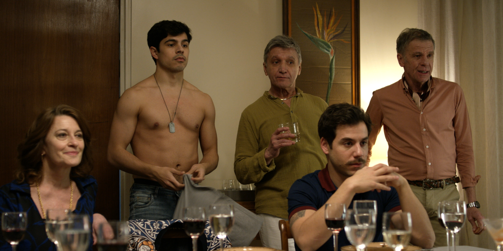 A man and woman sit at a dinner table while behind them stands a young shirtless man and two older, well-dressed men