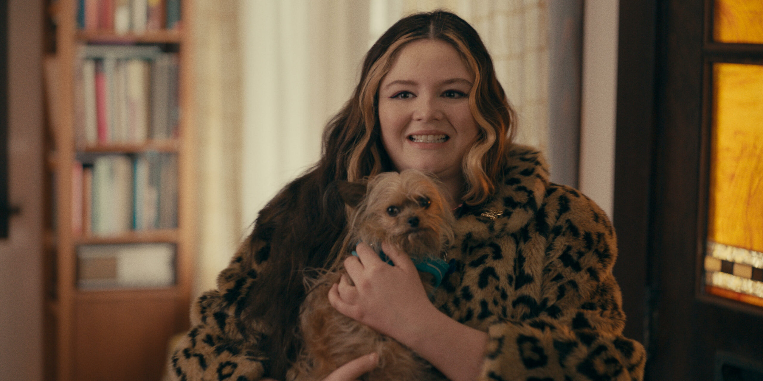 A woman wearing a faux fur coat smiles awkwardly as she holds a small dog