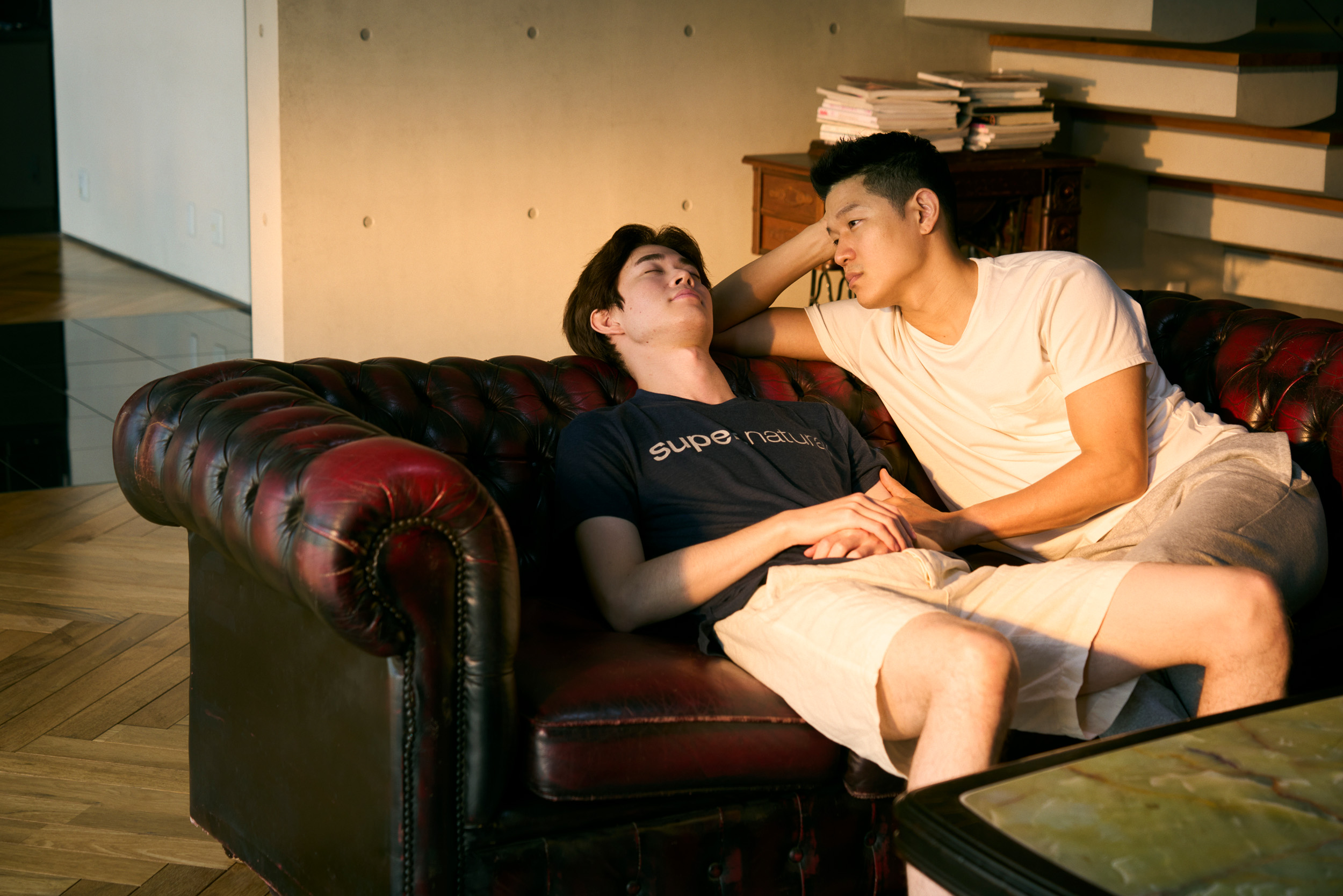 Two men sit on a sofa – one lays back with his eyes closed, the other rests his hand on his arm as he looks lovingly at him