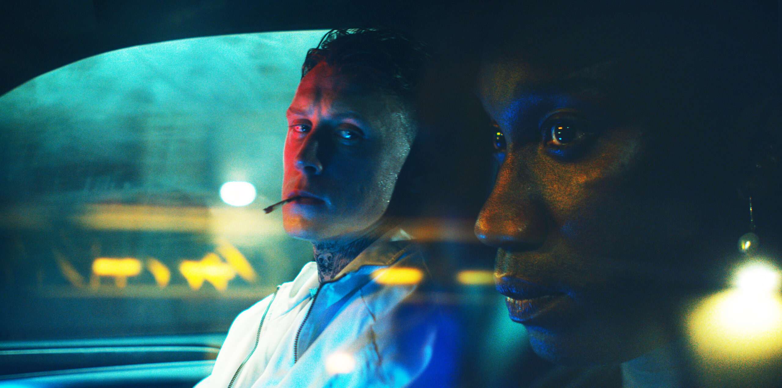 Two men sit in a car, bathed in neon lights – a white man with a cigarette in his mouth is looking at a black man, who looks out the window