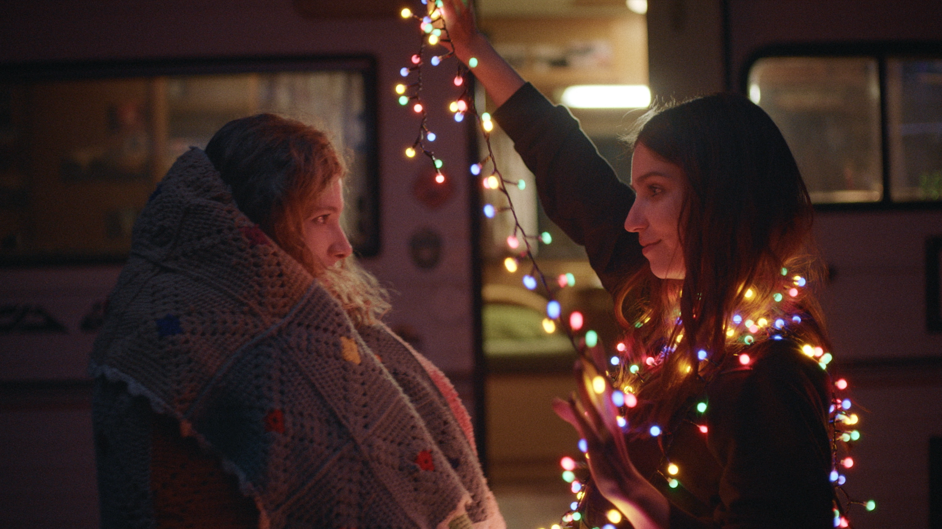 Two women smile at each other – one is wrapped in a blanker, the other a string of colourful fairy lights