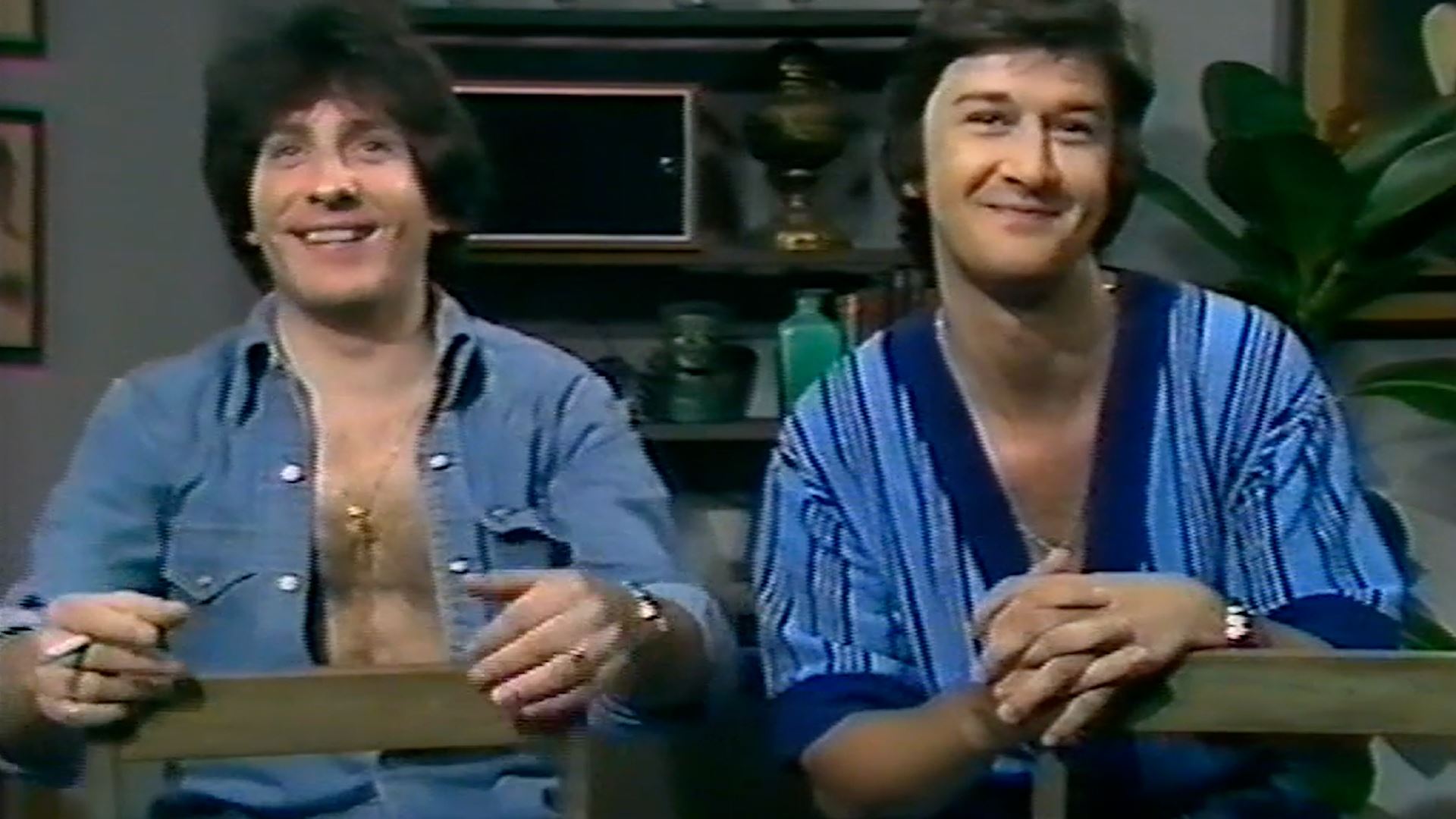Two masc people wearing open blue shirts smile at the camera. Based on the quality of the image, it looks like an old TV show.