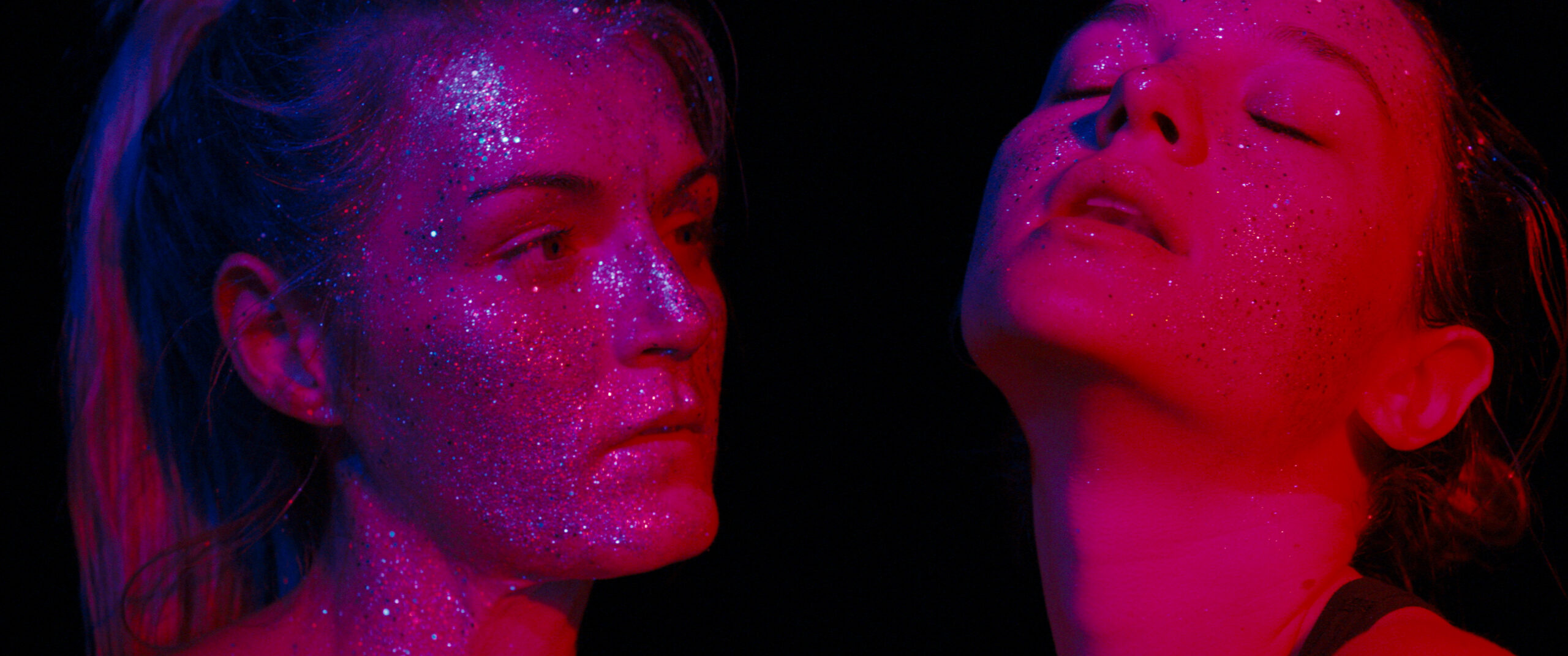 Two women dance under pink lighting, with glitter all over their faces