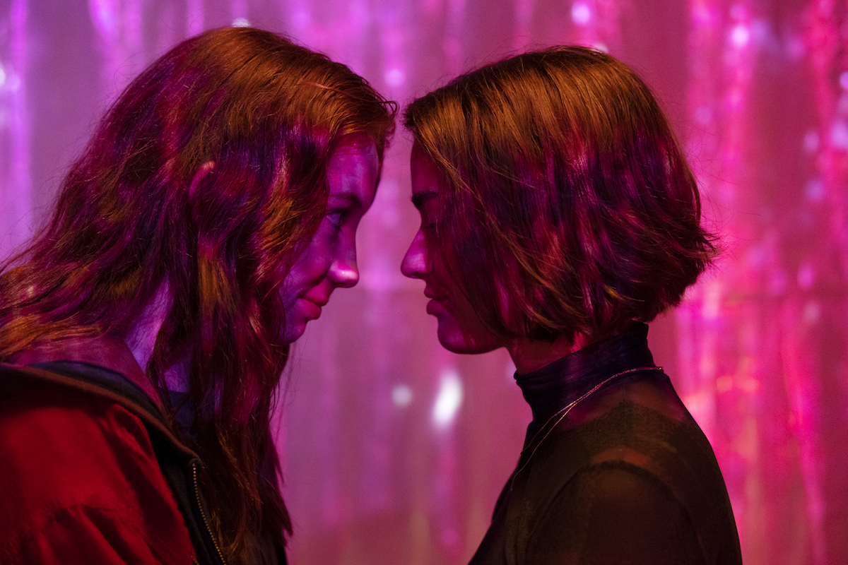 A pink-toned image, with two femme people staring at each other, foreheads nearly touching.