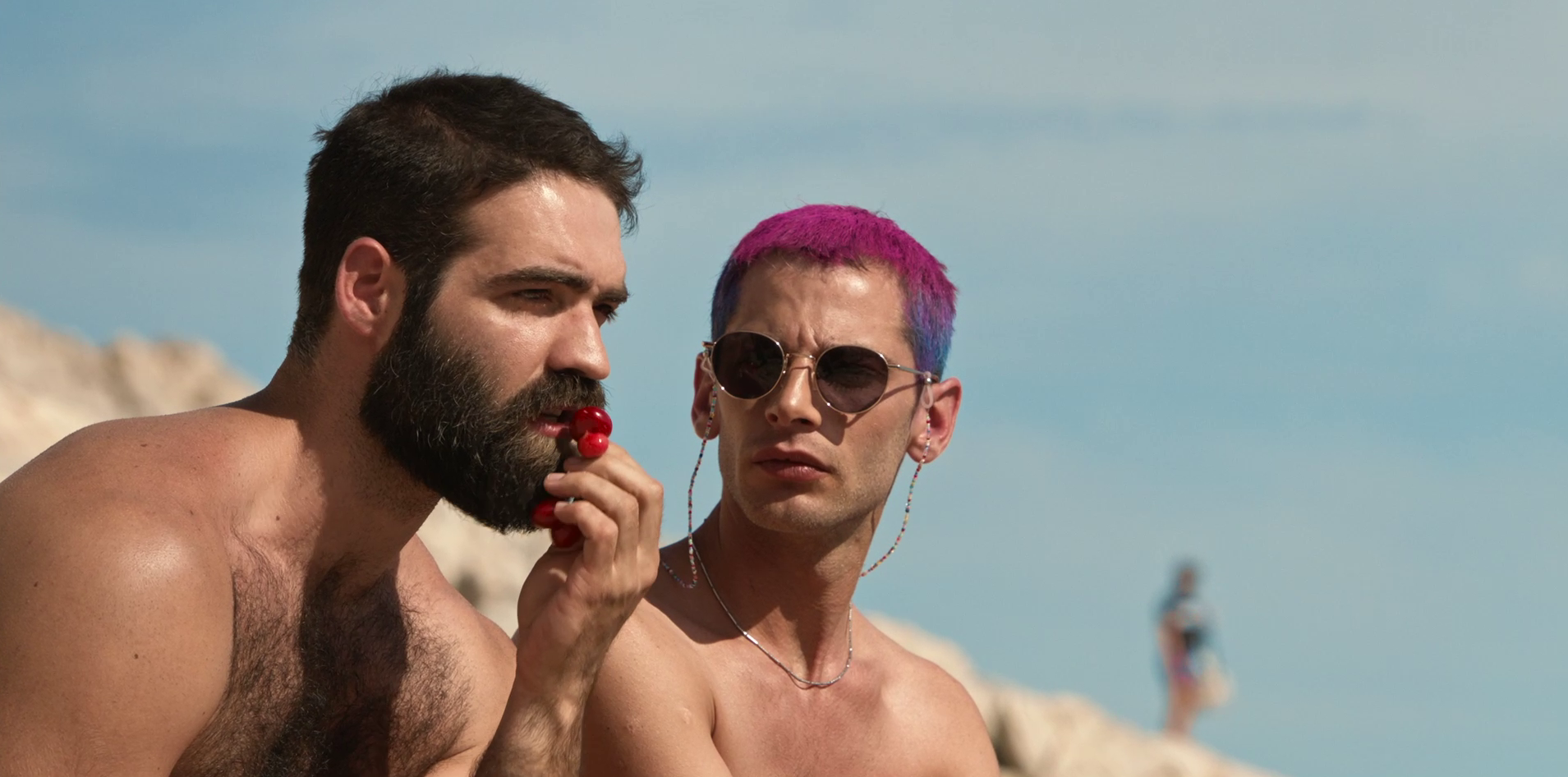 Two shirtless men sit on a beach: the bearded one is eating a cherry while the other, with purple and blue hair, is wearing sunglasses