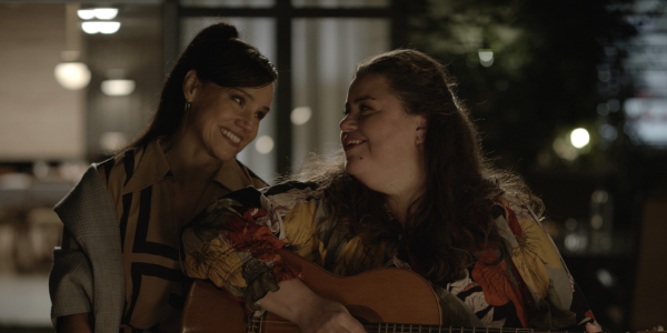 A still from the film 'Blue Lights'. Two people sitting next to each other, smiling, making eye contact. One is playing the guitar.