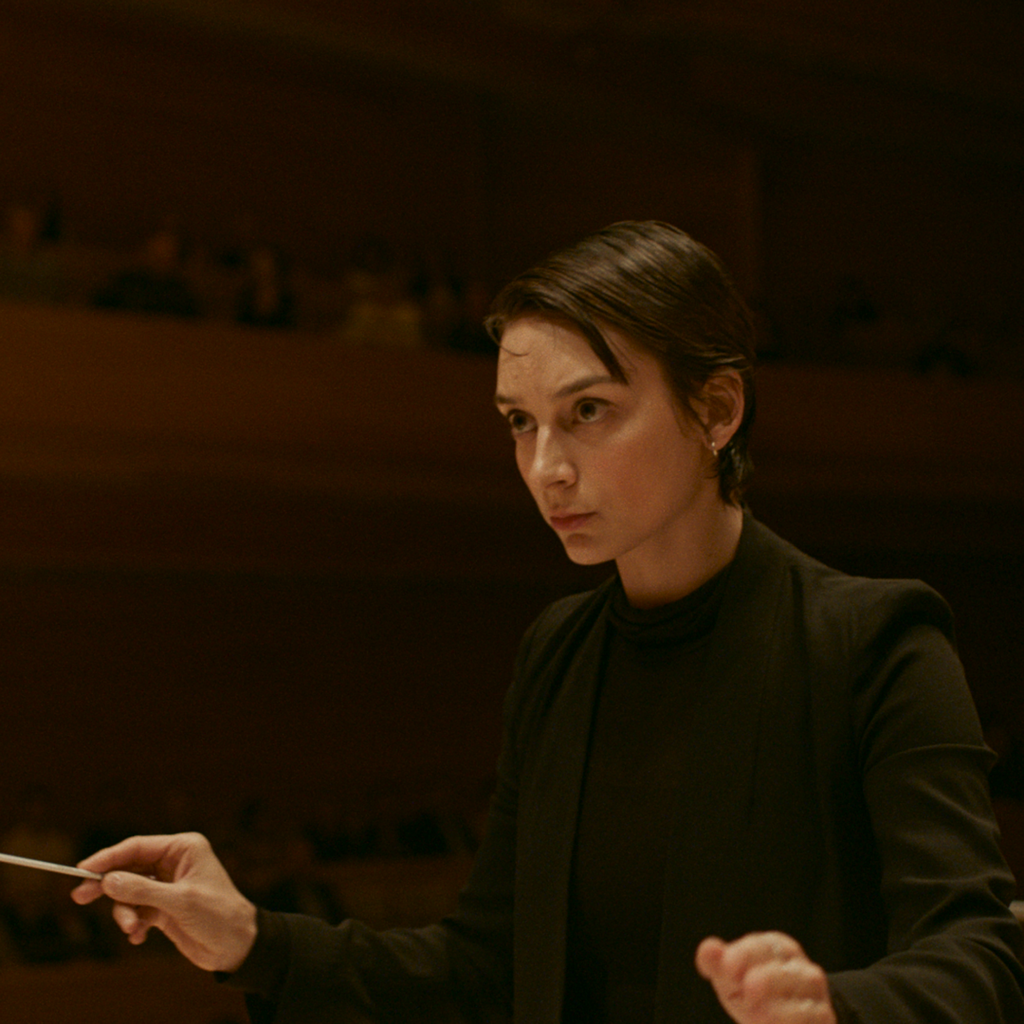 A woman stands holding a conductor's baton, an audience out of focus in the background.