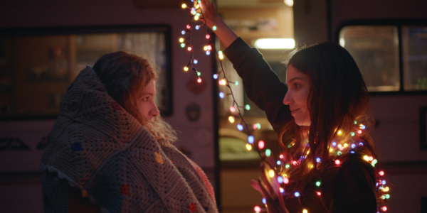 A still from the film 'Girls Don't Cry'. One person is wrapped in a knitted blanket, the other is wrapped in fairy lights.