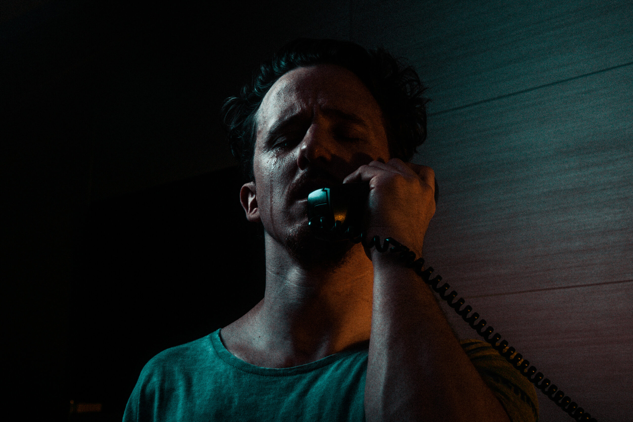 A man stands in darkness with an old-fashioned landline phone against his ear, a single tear running down his cheek.