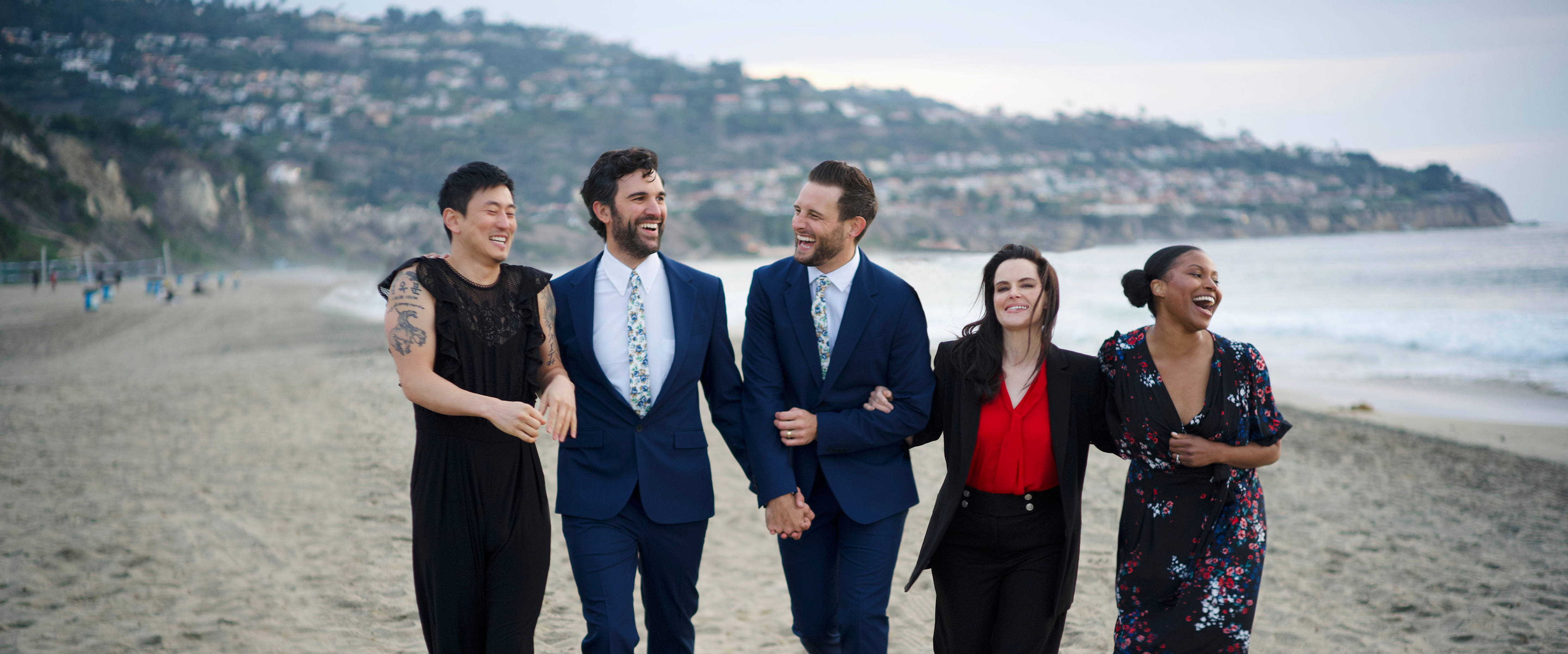 Five people walk arm-in-arm along a beach, smiling and laughing.