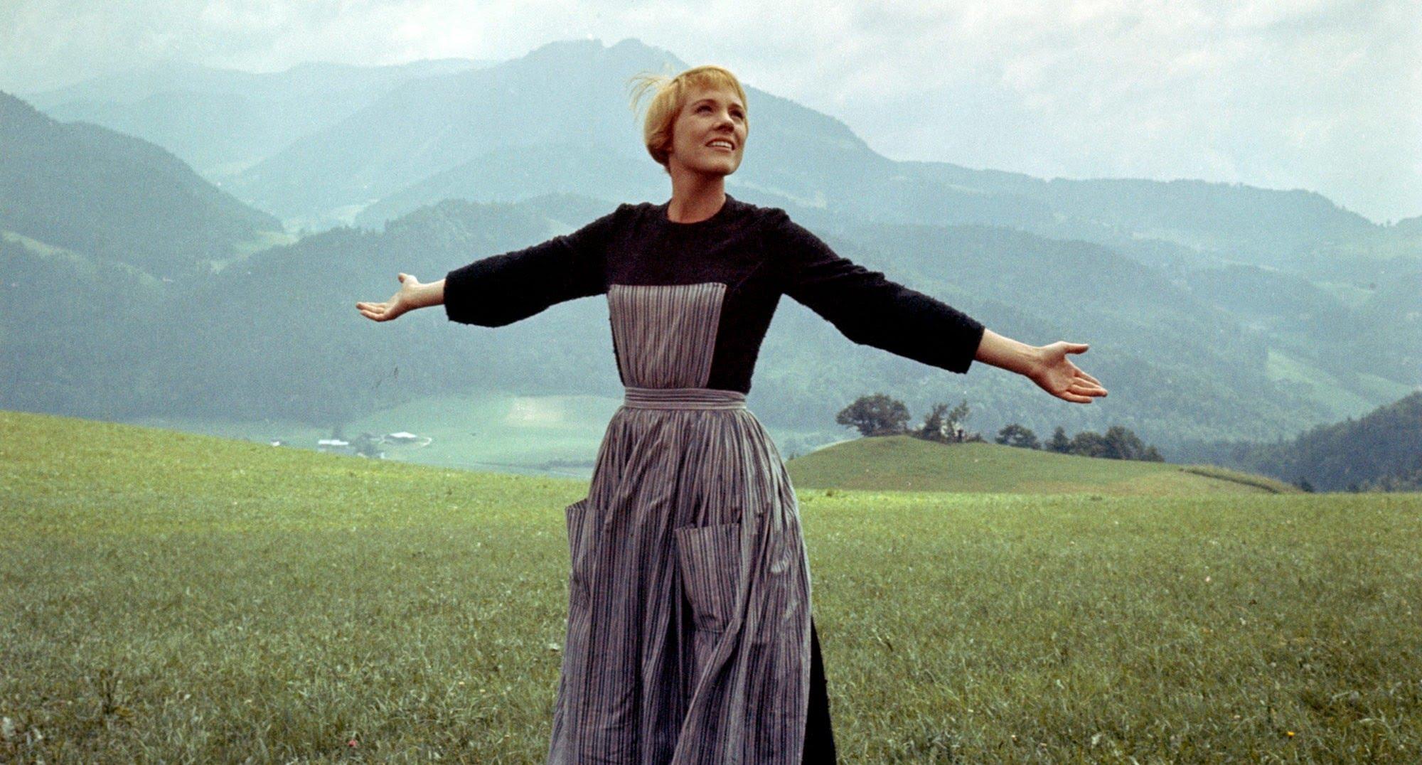 A woman stands on a grassy hill with the Alps in the background, arms raised as if she's just finished singing.