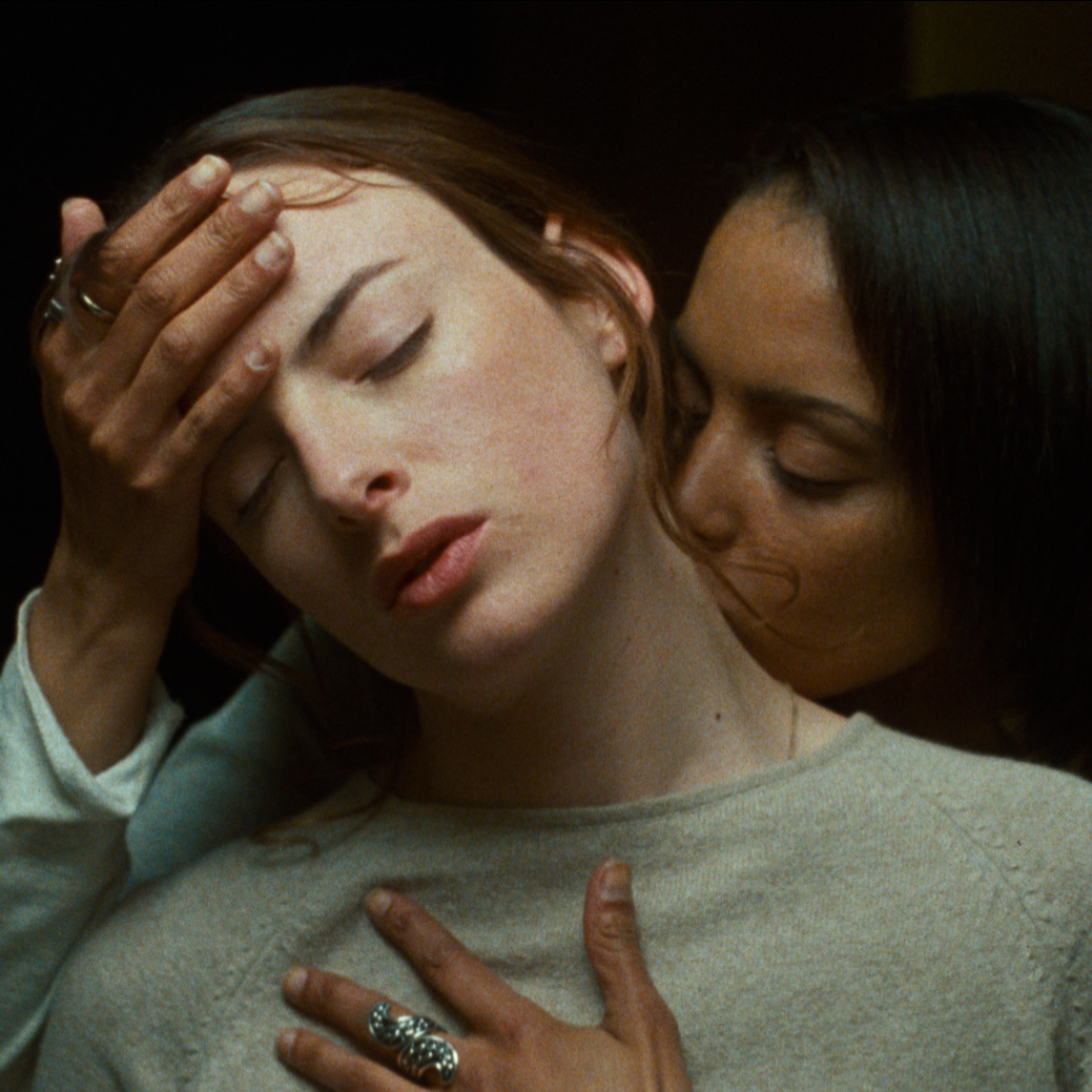 A woman kisses another woman from behind, her hands resting on her partner's head and chest.