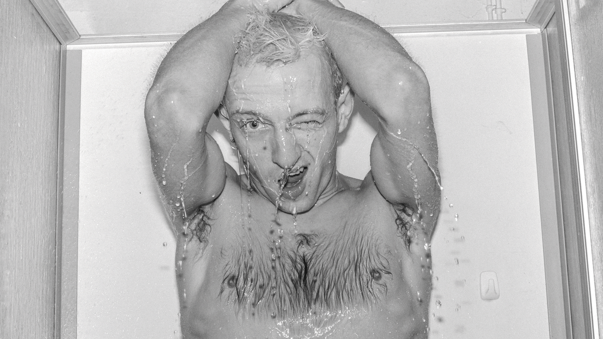 A black-and-white photo of a shirtless man winking at the camera from a shower.