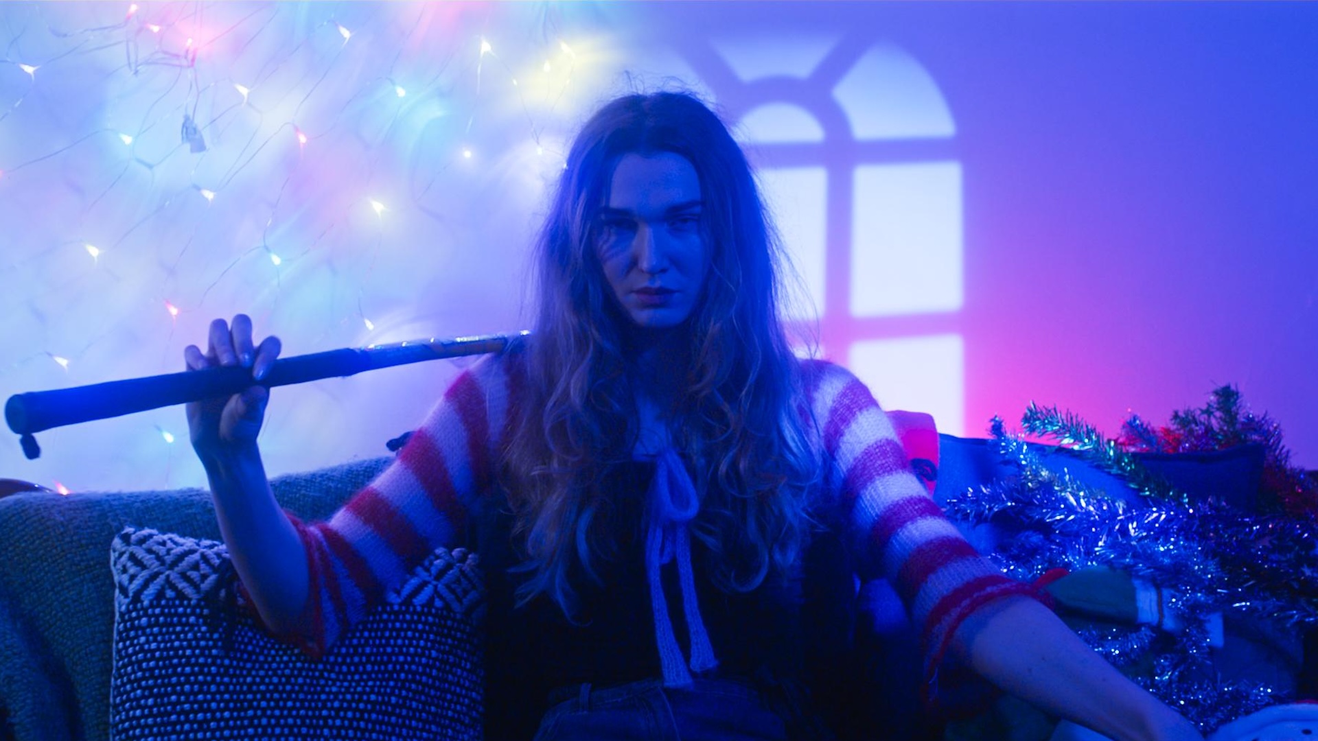 A women sits in a couch, poised with a weapon over her shoulder, lit by Christmas fairy lights