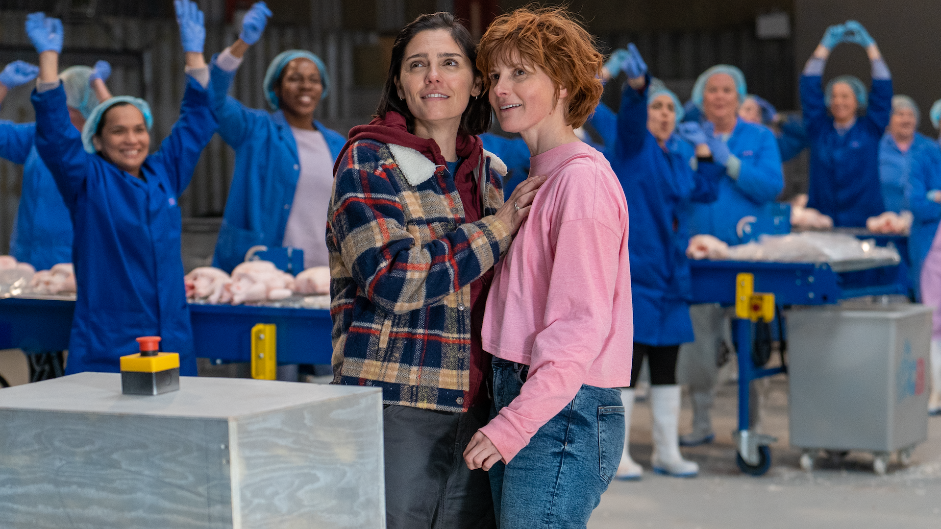 In the background factory workers celebrate, while two smiling women stand in front of them holding each other