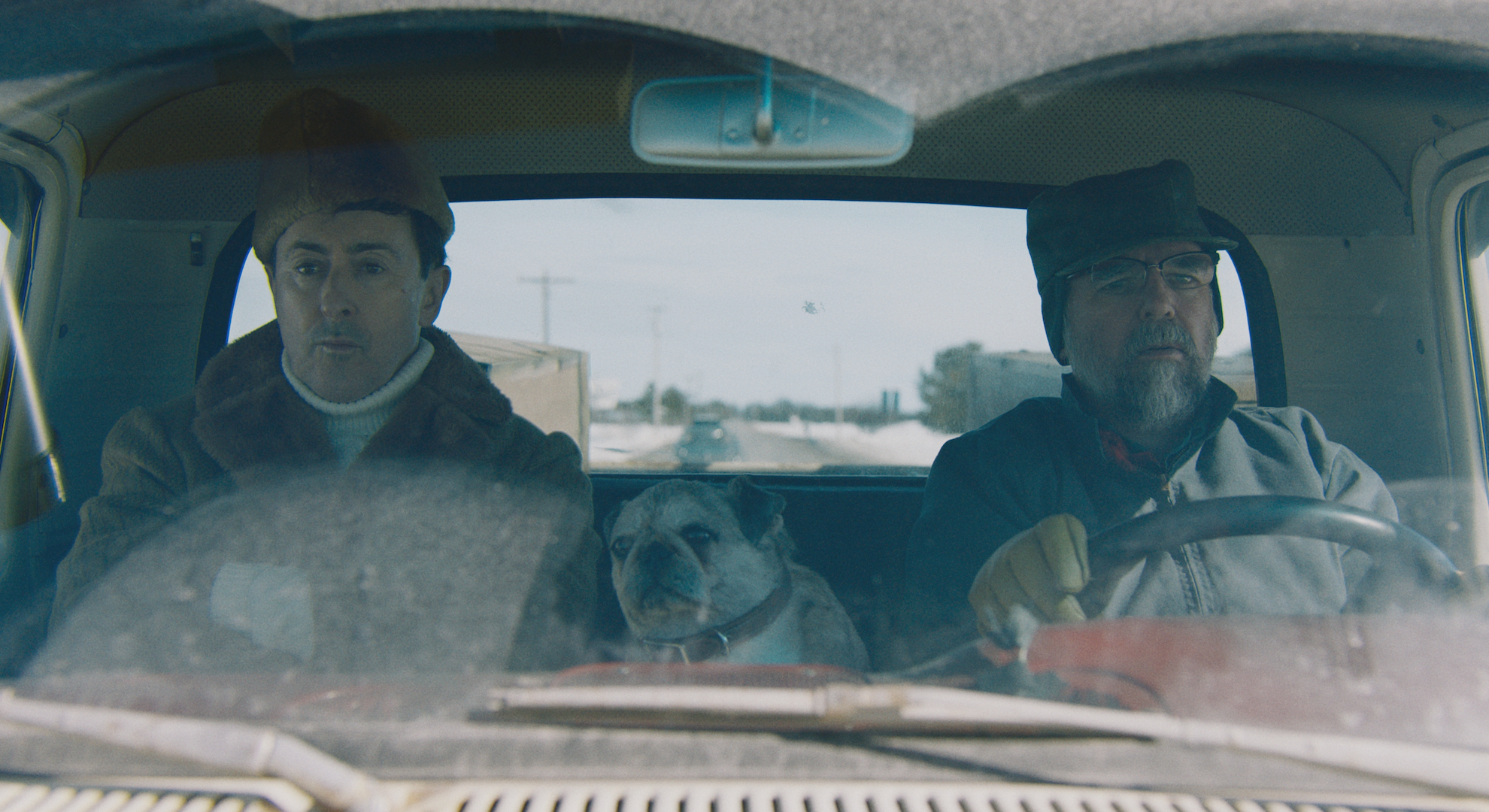 Two men sit in a car staring straight ahead, a taxidermied bug sitting between them.