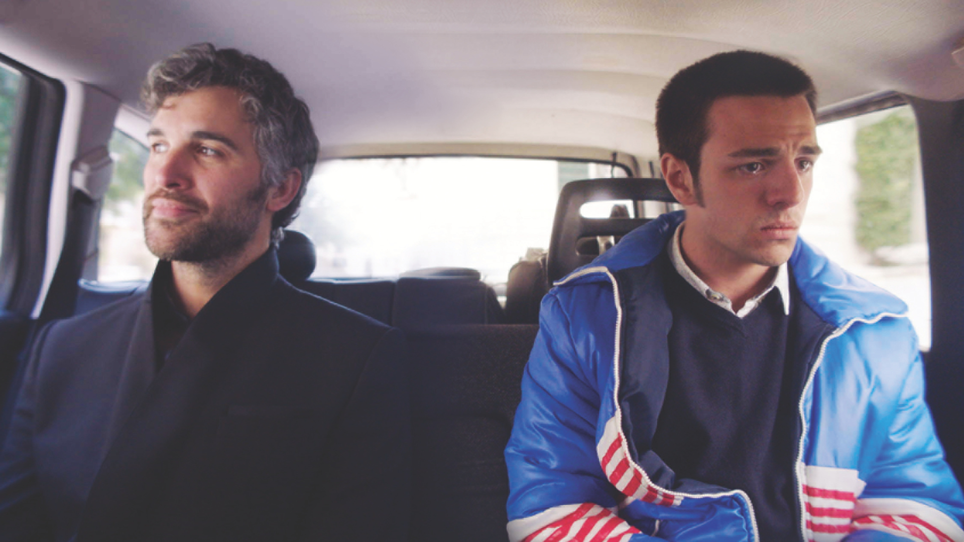 Two men sit in the back of a car: one is older and smiling slightly, the other younger and looking distraught.