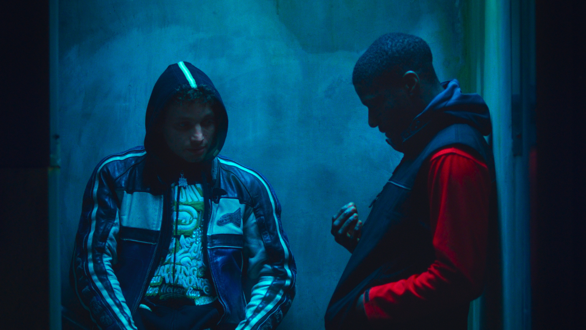 Two men lean against a wall in shadows, one has his hood up while the other looks at something he's holding