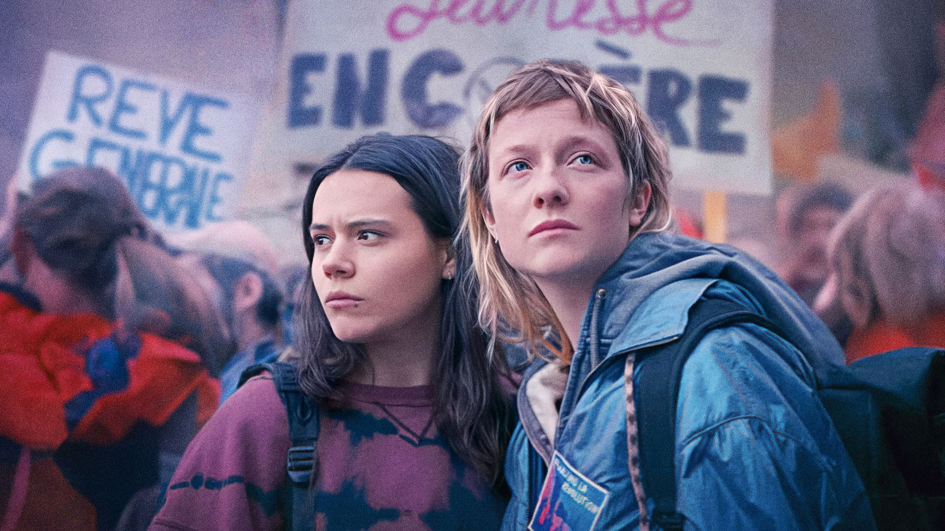 Two teenage girls stand amidst a protest, with people holding placards