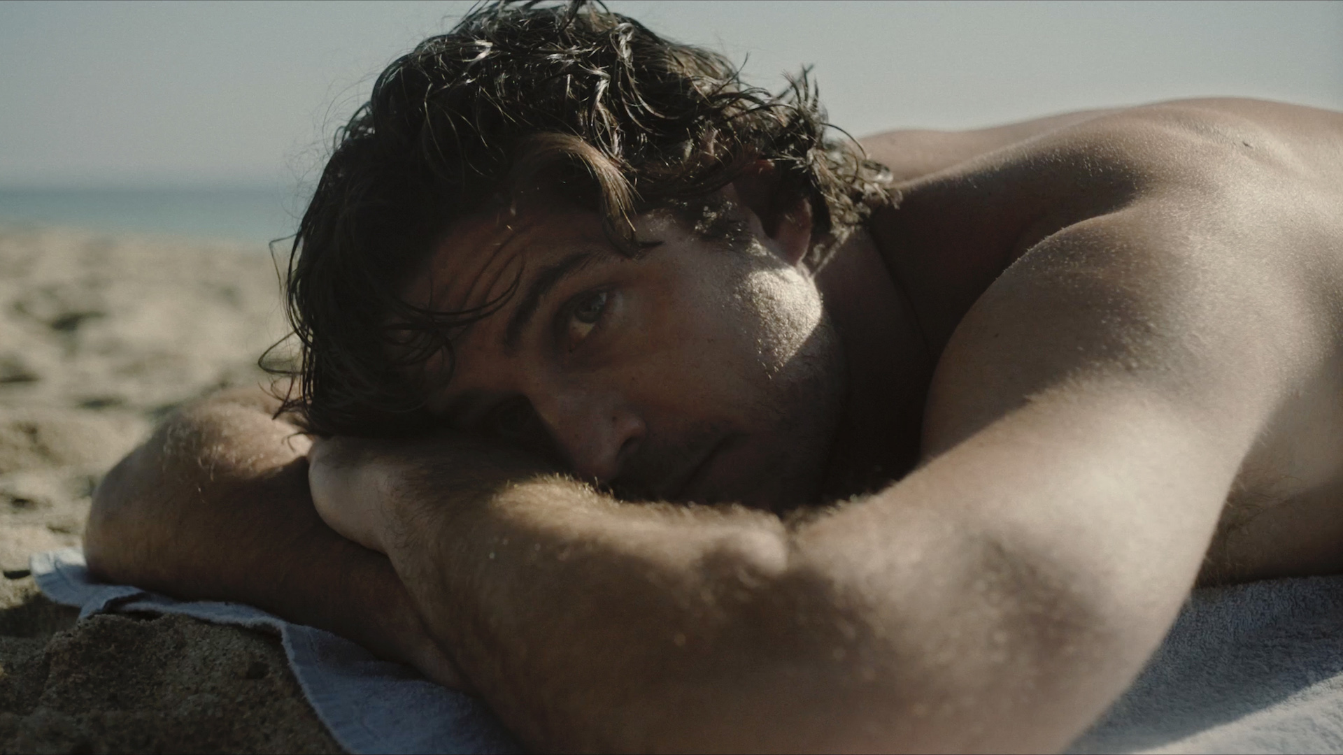 A shirtless man lies on a beach, resting his head on his arms.