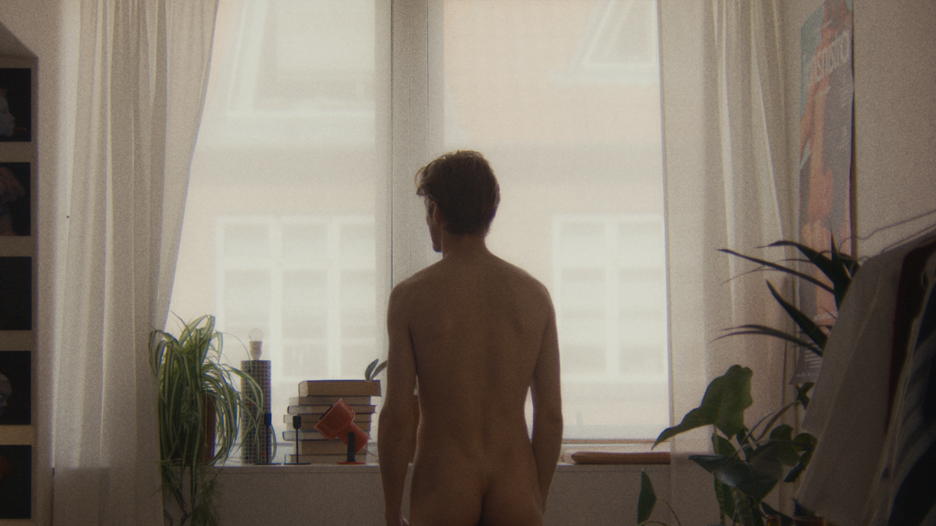 A naked man looks out a window, his bum on display