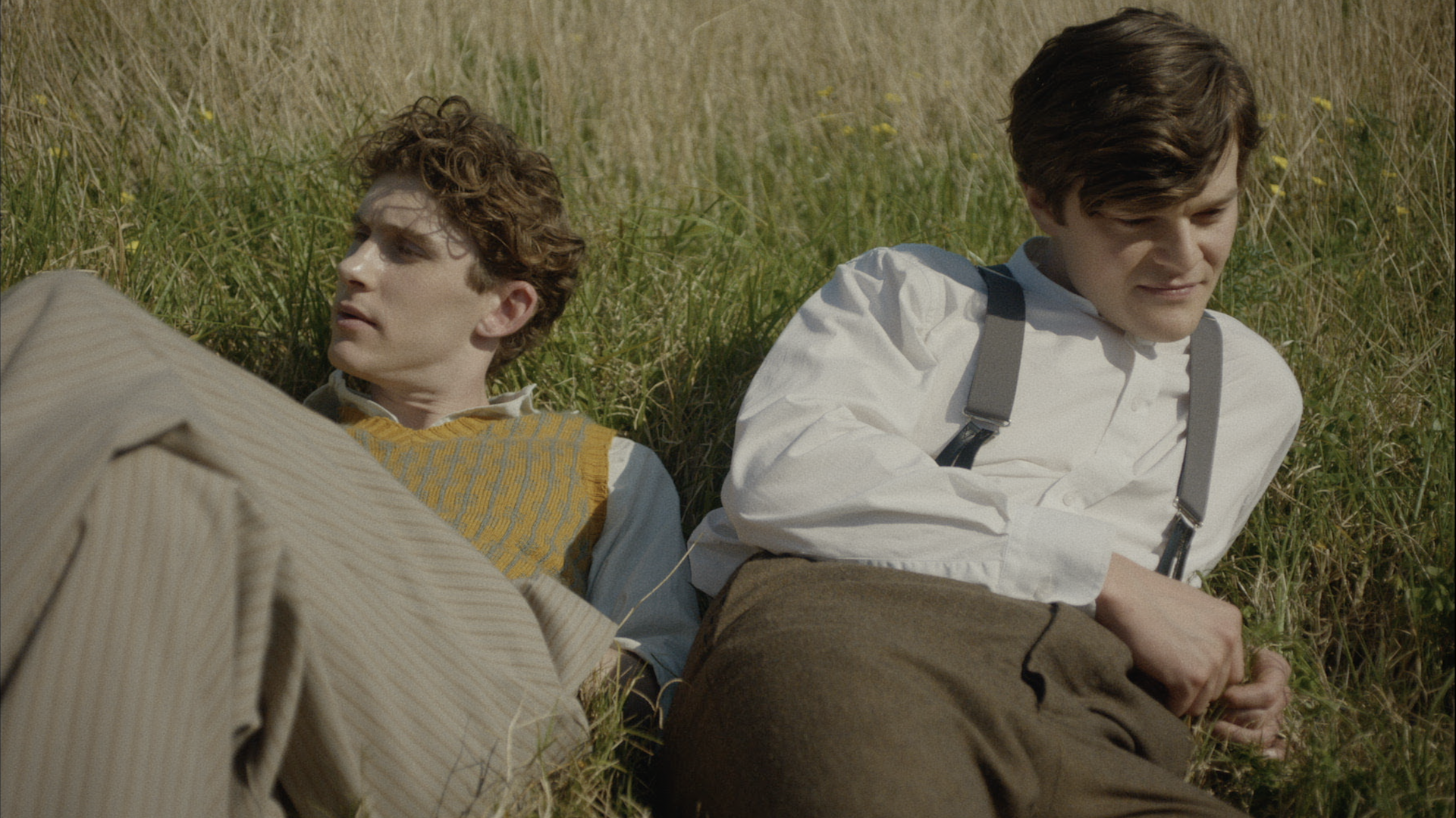 Two men in 1920s period clothing lounge in the grass together.