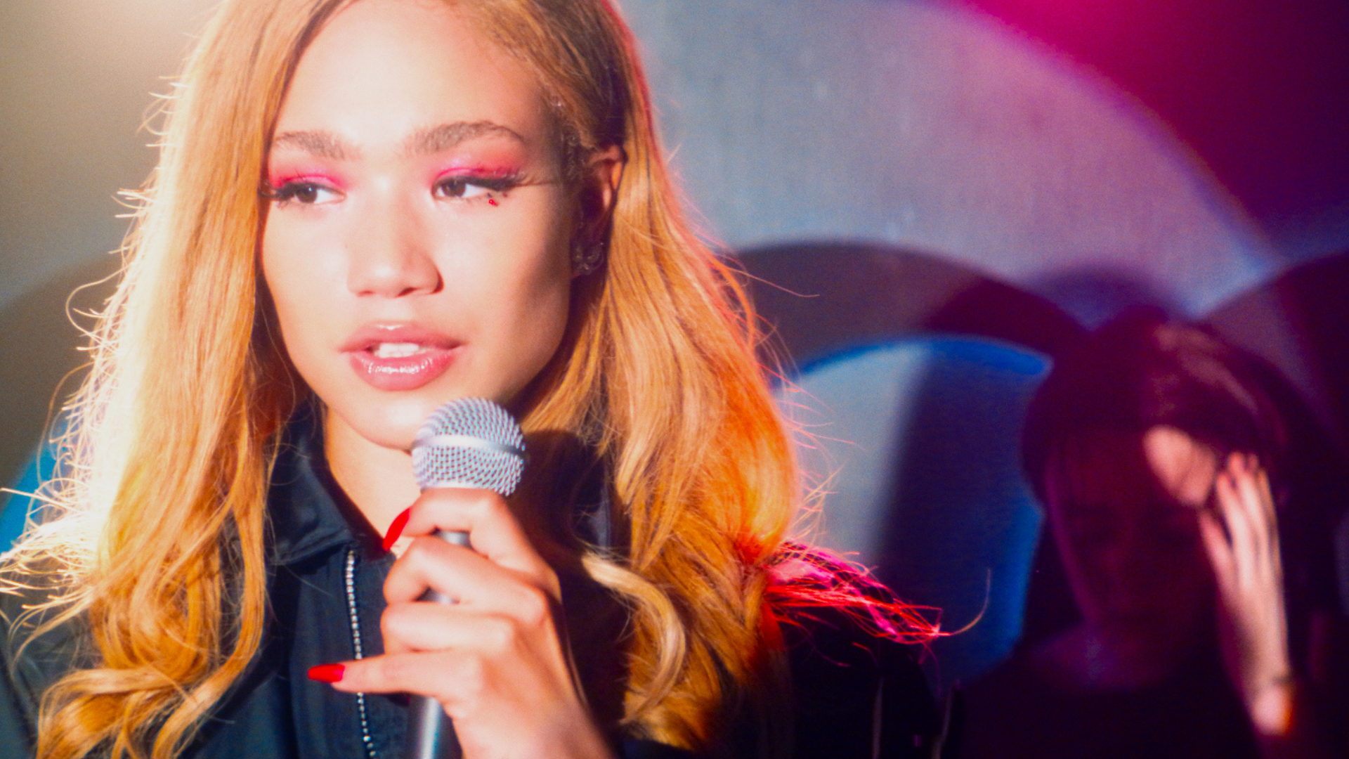 In the foreground, a young woman lit by a spotlight holds a microphone, while in the background another woman is bathed in shadows