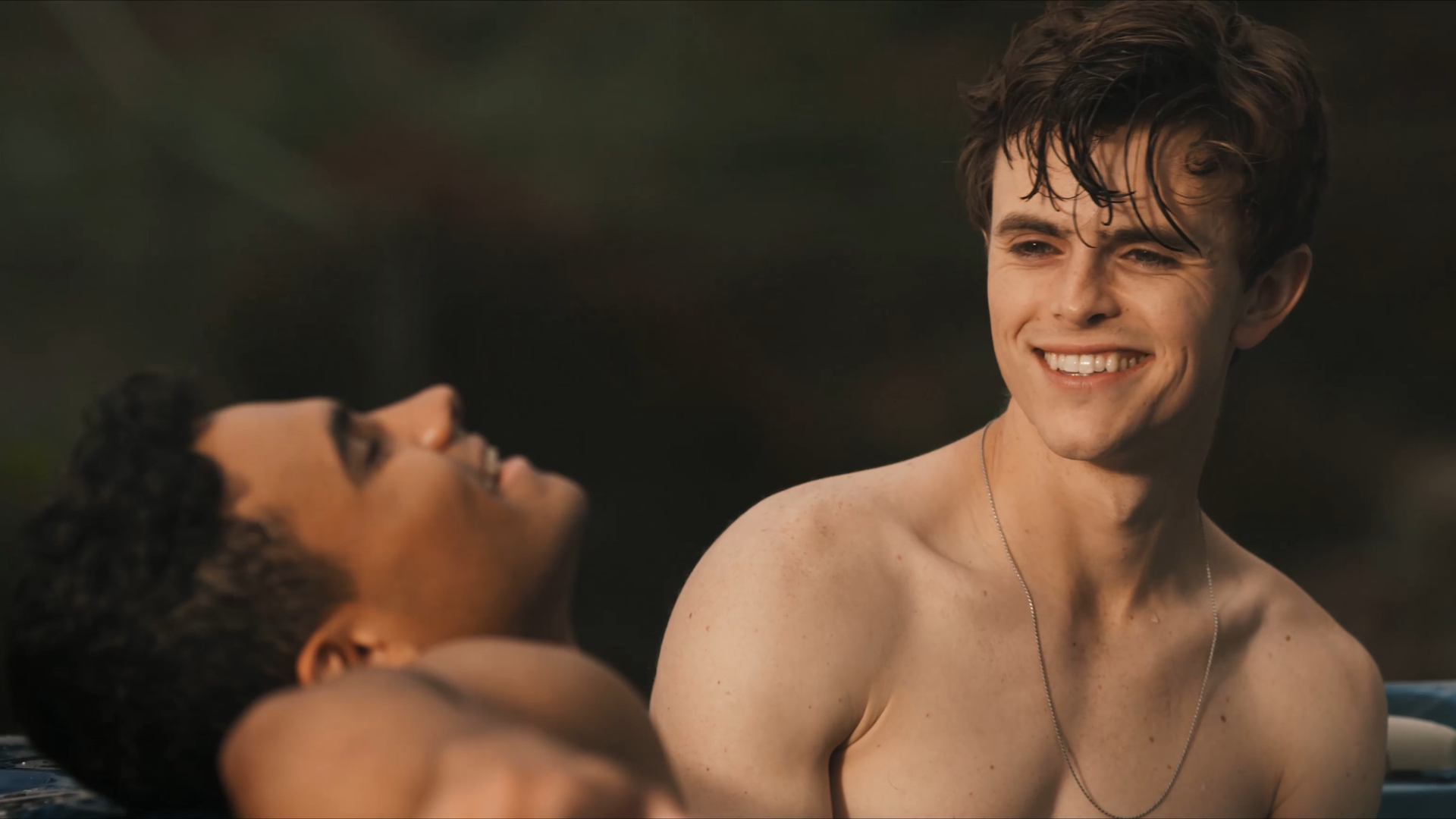 A young shirtless man smiles at another who is leaning back, looking upwards