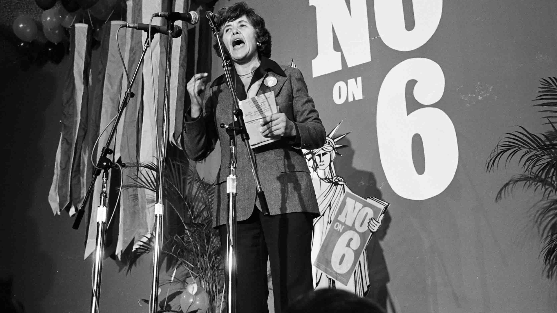 A black-and-white archival photo of a woman from the 1970s speaking into a microphone in front of the words "No on 6"