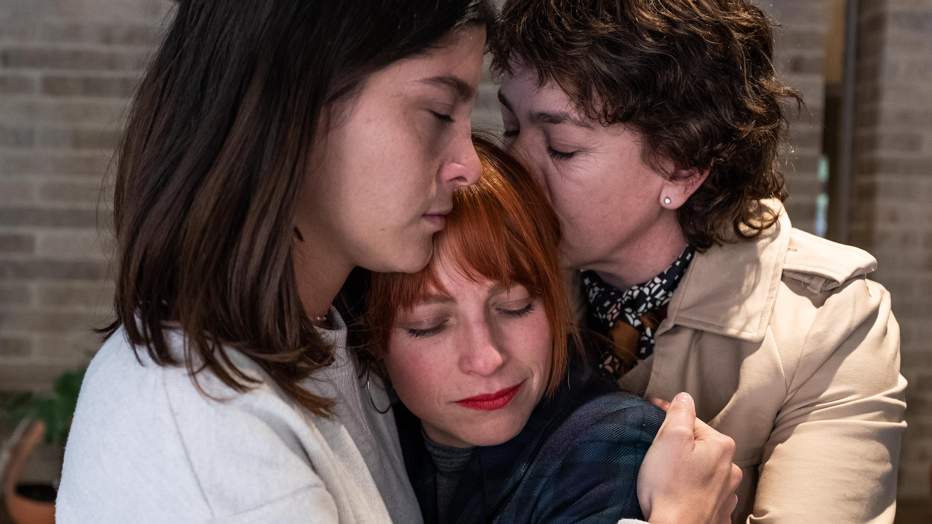 Three women hug each other, the one on the right kissing the head of the one in the centre