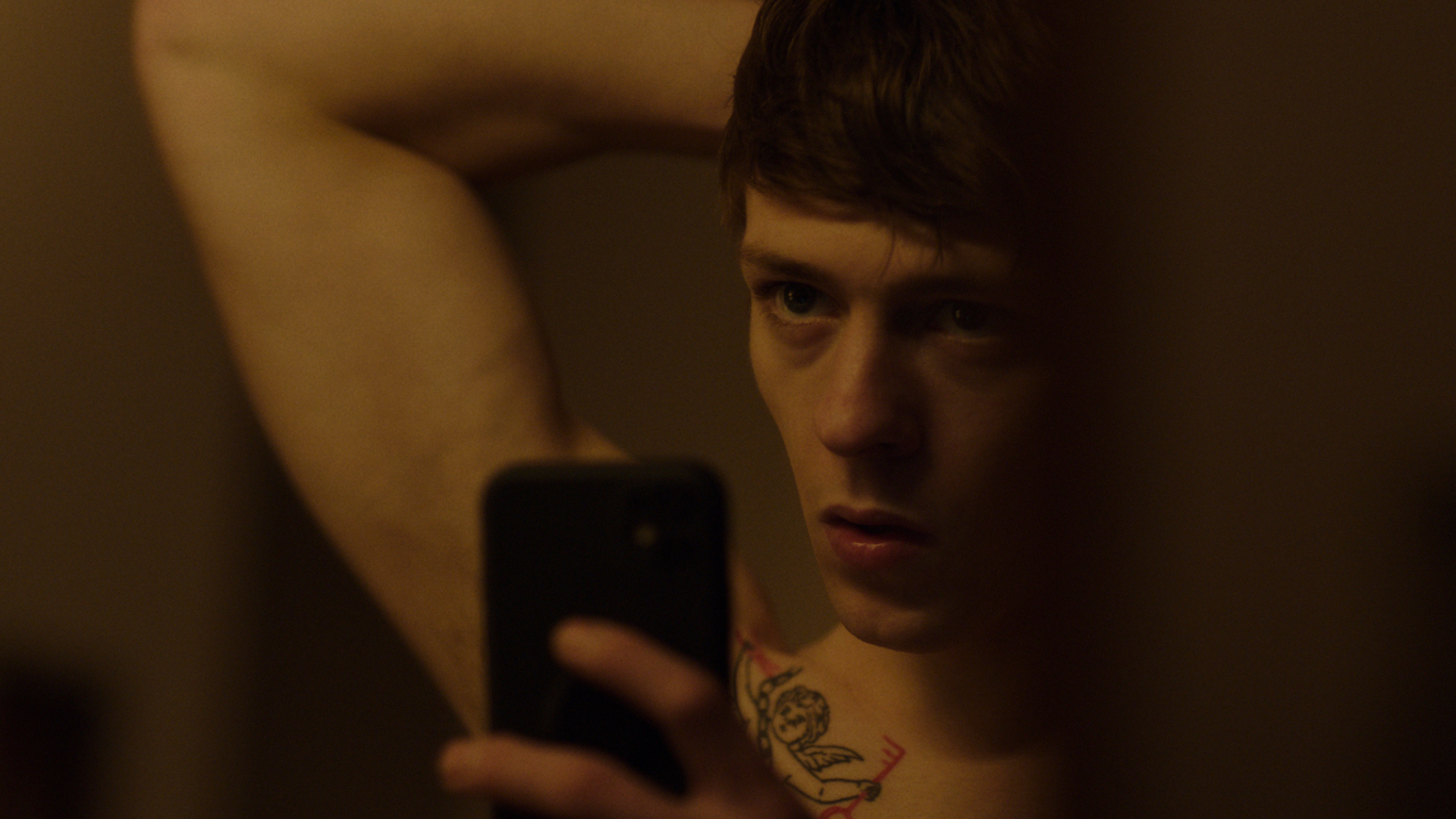 A young shirtless man raise his arm behind his head as he takes a selfie in a mirror