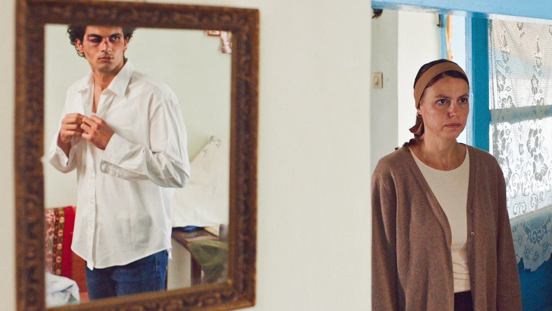 A woman stands looking at a man reflected in a mirror, who has bruising on his face