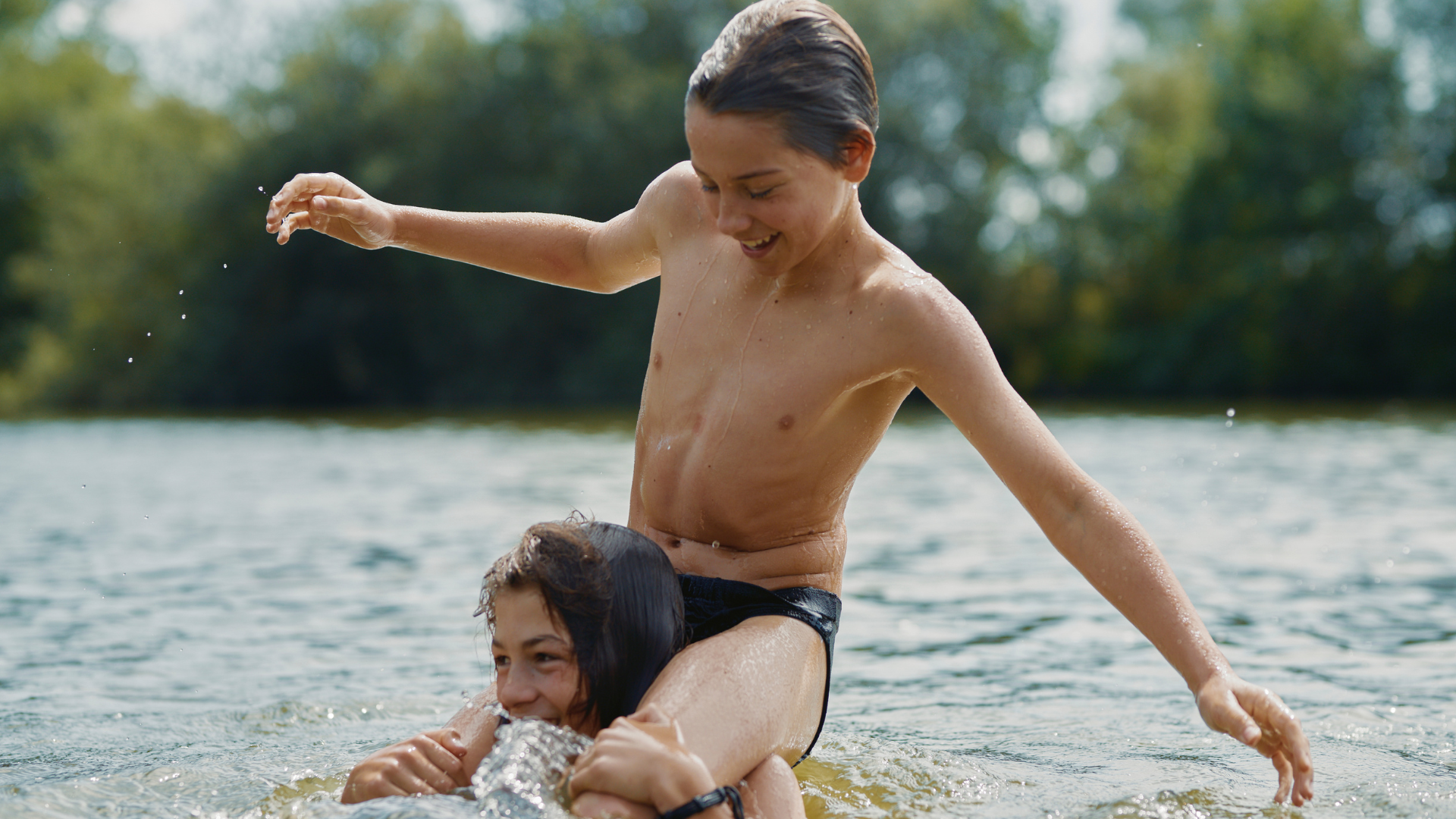 Two young, smiling teenage boys play in a lake, one sitting on the other's shoulders.