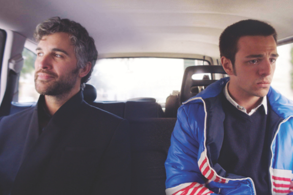 Two men sitting together in the back seat of a car. Both looking out the window. One of them looks sad, the other looks happy.