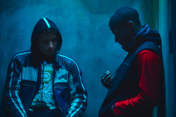 Two men in dim lit corner with faces hidden, one is wearing a jacket with hood on and the other is facing down at hands