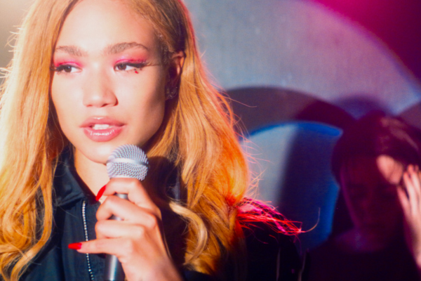 Female holding microphone and standing in spotlight. She has bright make up and dyed orange hair