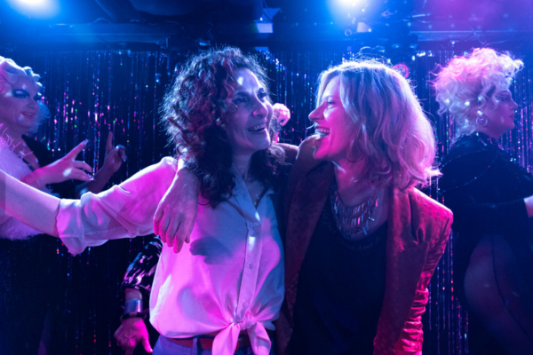 Two women embracing on stage, singing and surrounded by drag queens