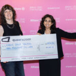 Two people in black clothing standing in front of a media wall carrying a giant novelty check.